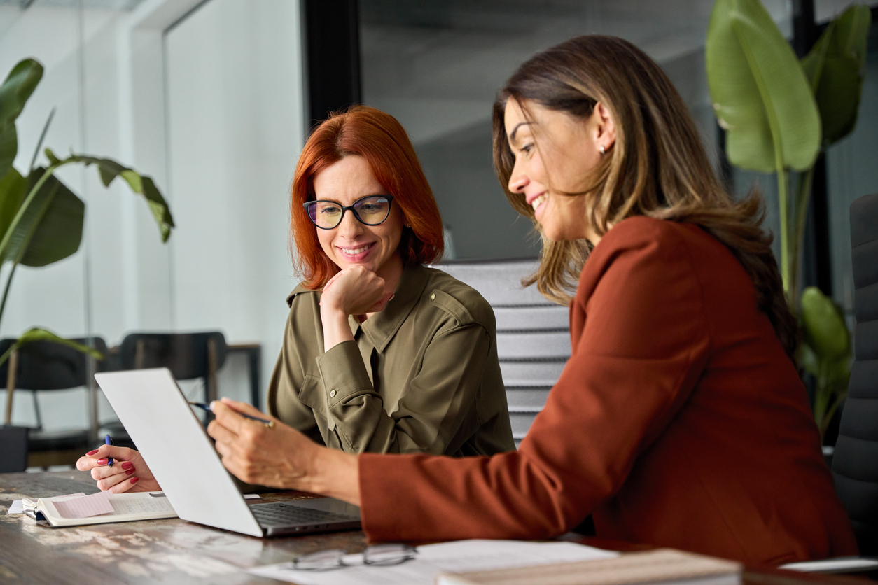 Two ha[[y busy female coworkers working together using computer at desk in office.